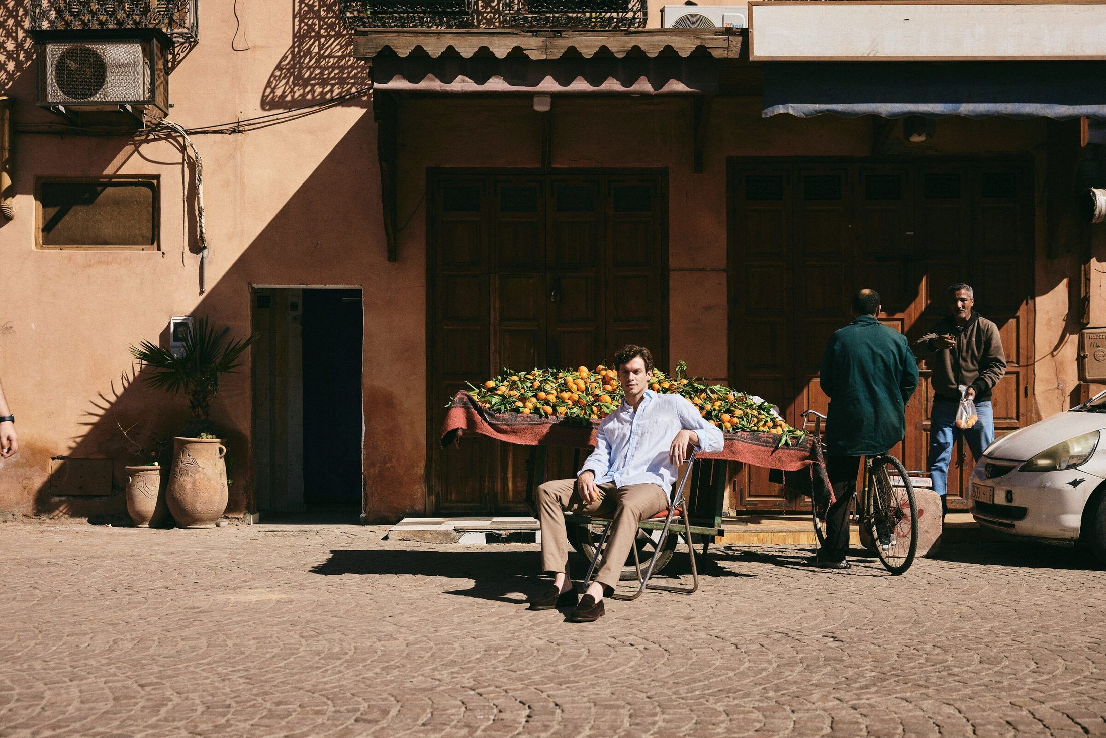 Male model seated beside a cart of oranges in front of an ochre wall in a Marrakech street – Emporio Models Marrakech