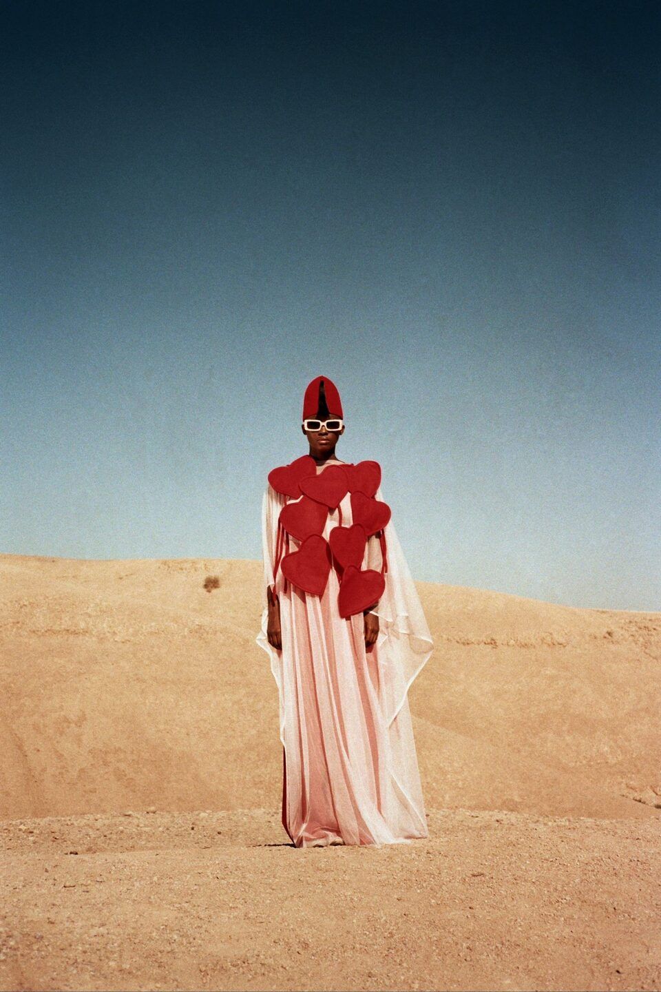 Mannequin en tenue drapée rouge et blanche, seule dans un désert de sable sous un ciel bleu profond – Emporio Models Marrakech
