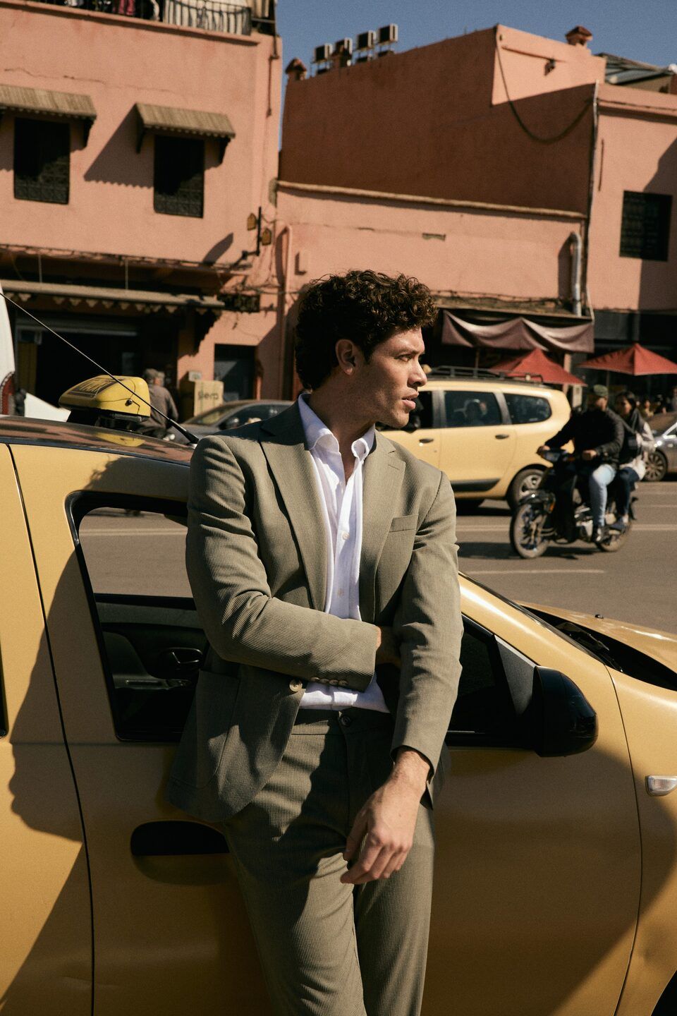 Mannequin homme en costume olive appuyé sur un taxi dans une rue de Marrakech bordée de murs roses – Emporio Models Marrakech