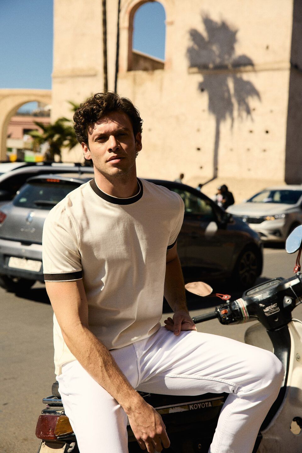 Mannequin homme en t-shirt crème et pantalon blanc sur un scooter dans une rue de Marrakech bordée de palmiers et de vieux murs – Emporio Models Marrakech